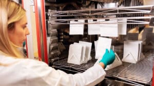 A scientist wearing green gloves handles paper packaging samples on shelves in a laboratory chamber.