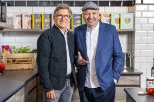 Johann Lafer wearing a grey cap and blue jacket beside a man in glasses in an EDEKA kitchen