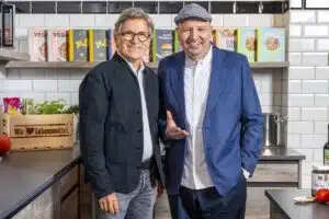Johann Lafer wearing a grey cap and blue jacket beside a man in glasses in an EDEKA kitchen