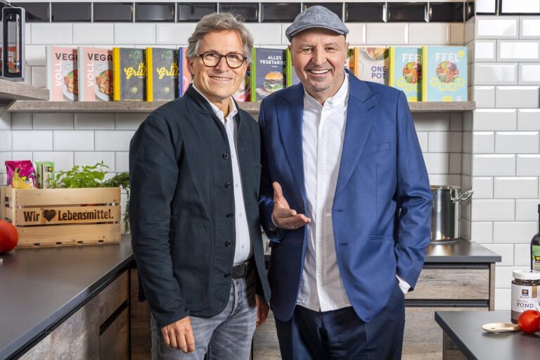 Johann Lafer wearing a grey cap and blue jacket beside a man in glasses in an EDEKA kitchen