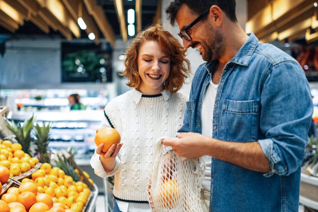 Two shoppers selecting oranges in a supermarket produce aisle.