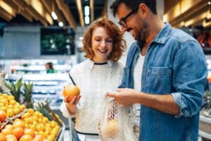 Two shoppers selecting oranges in a supermarket produce aisle.