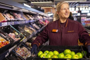 A Sainsbury’s colleague holding a crate of green apples beside a produce display.