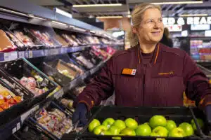 A Sainsbury’s colleague holding a crate of green apples beside a produce display.