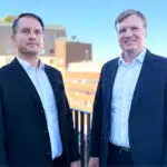 Two Scandi Standard executives in dark jackets standing outdoors beside a railing.