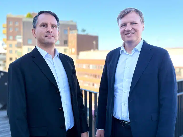 Two Scandi Standard executives in dark jackets standing outdoors beside a railing.