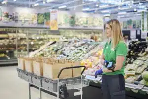 Supermarket worker with a wearable scanner beside a trolley holding paper grocery bags