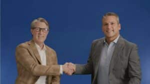 Two men in jackets shake hands in front of a blue background.