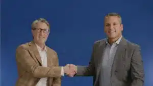 Two men in jackets shake hands in front of a blue background.